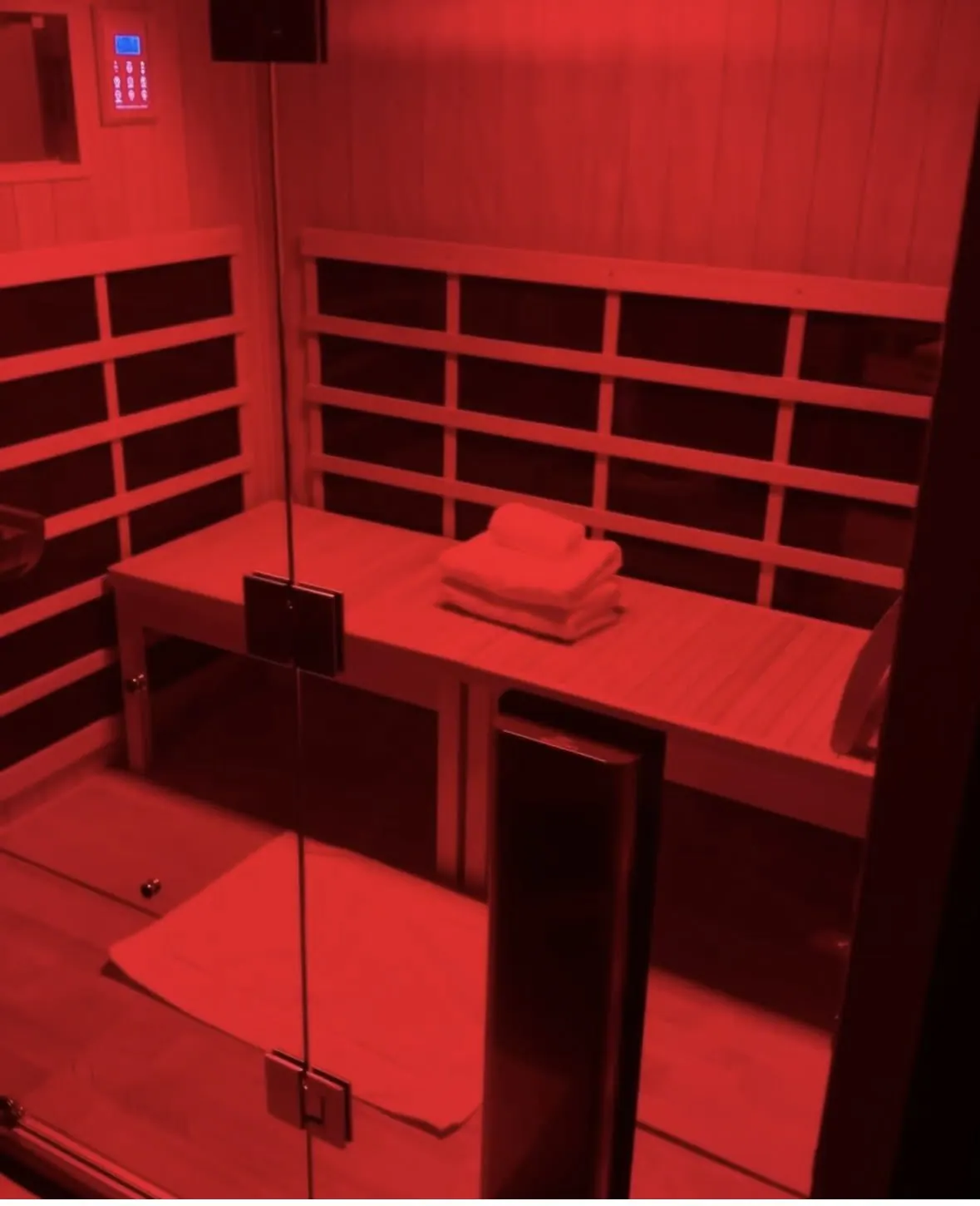 A red-colored sauna room with wooden slats on the walls. There are folded towels placed on a wooden bench inside the sauna and a mirror is reflecting part of the room.